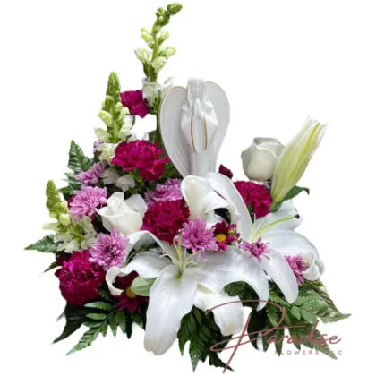 Angelic Blessings floral arrangement featuring white roses, snapdragons, purple carnations, purple cushion, white lilies, and an angel keepsake sculpture.