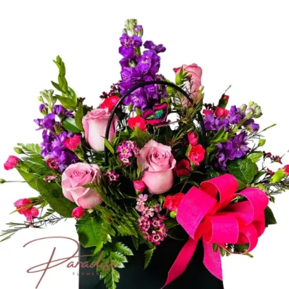 Enchanting Blossom Handbag floral arrangement featuring purple stocks, pink roses, hot pink mini carnations, waxflower accents, and rich greenery in a chic, waterproof cardboard handbag.