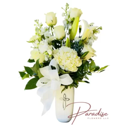 Peaceful Tribute Arrangement with white roses, lilies, stocks, and mini carnations in a white ceramic vase with a silver cross and a white bow.