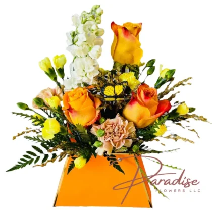A vibrant Little Harvest Charm fall flower arrangement featuring orange roses, white stock, and yellow mini carnations in a modern orange vase, perfect for autumn.