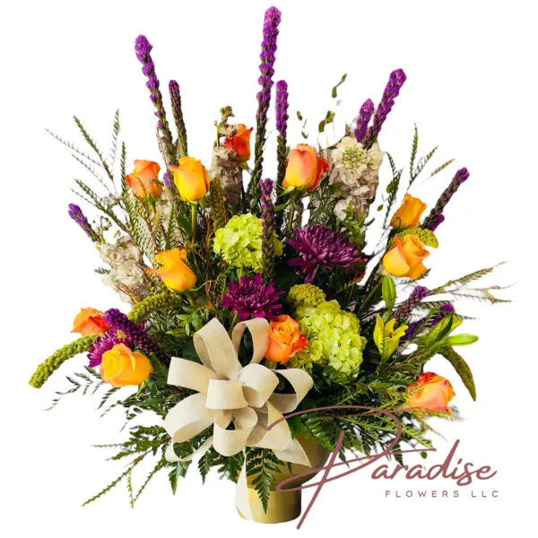 A vibrant floral arrangement featuring orange roses, purple mums, and green hydrangeas, accented with tall purple liatris, yellow lilies, and various fall foliage, set in a gold metallic vase with a beige bow.