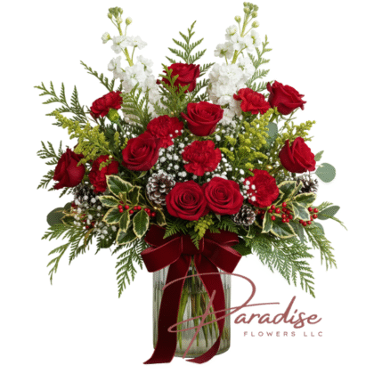 The Christmas Spirit Bouquet: A traditional holiday floral arrangement in a clear glass vase featuring red roses, red carnations, white stock, rustic pinecones, seasonal greens, holly, and a large red velvet bow.